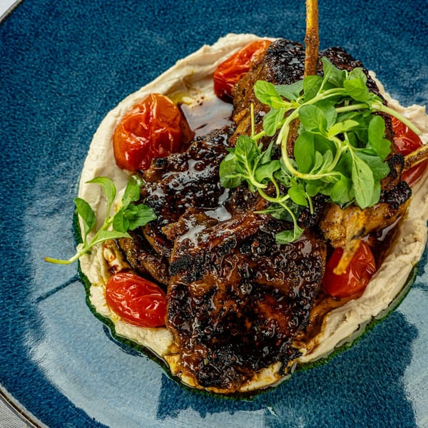 Glazed meat skewers with roasted tomatoes and greens on a bed of hummus, served on a blue plate.