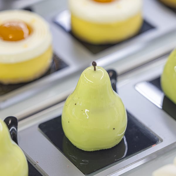 A vibrant green pear-shaped pastry with a glossy glaze sits on a black plate, with other pastries blurred in the background.