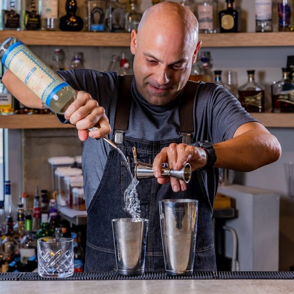 A bald bartender in a denim apron pours liquid from a bottle into a jigger and then a shaker.