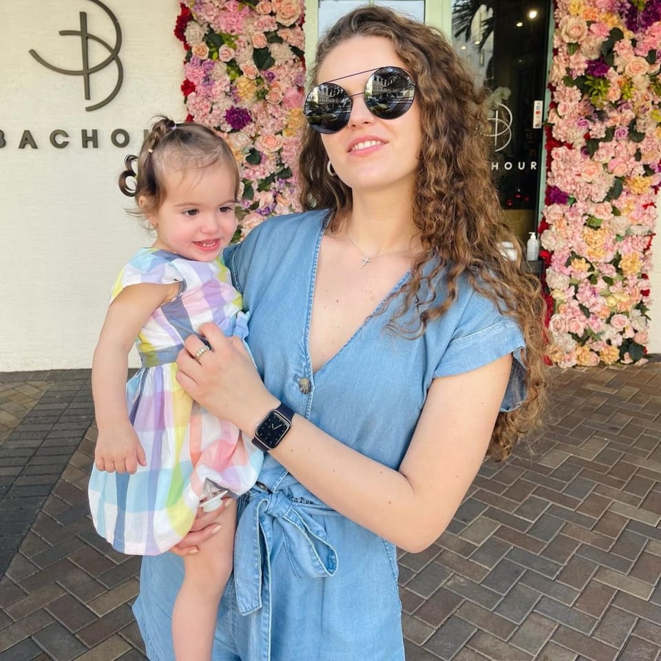 A woman in sunglasses holding a baby in front of Bachour bakery's floral wall.