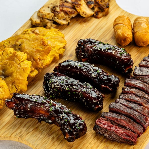 A wooden board with grilled chicken, ribs, fried plantains, and two fried pastries.
