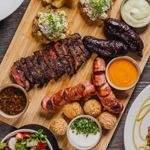 A wooden board with sliced steak, sausages, blood sausage, small fried balls, and various dipping sauces.