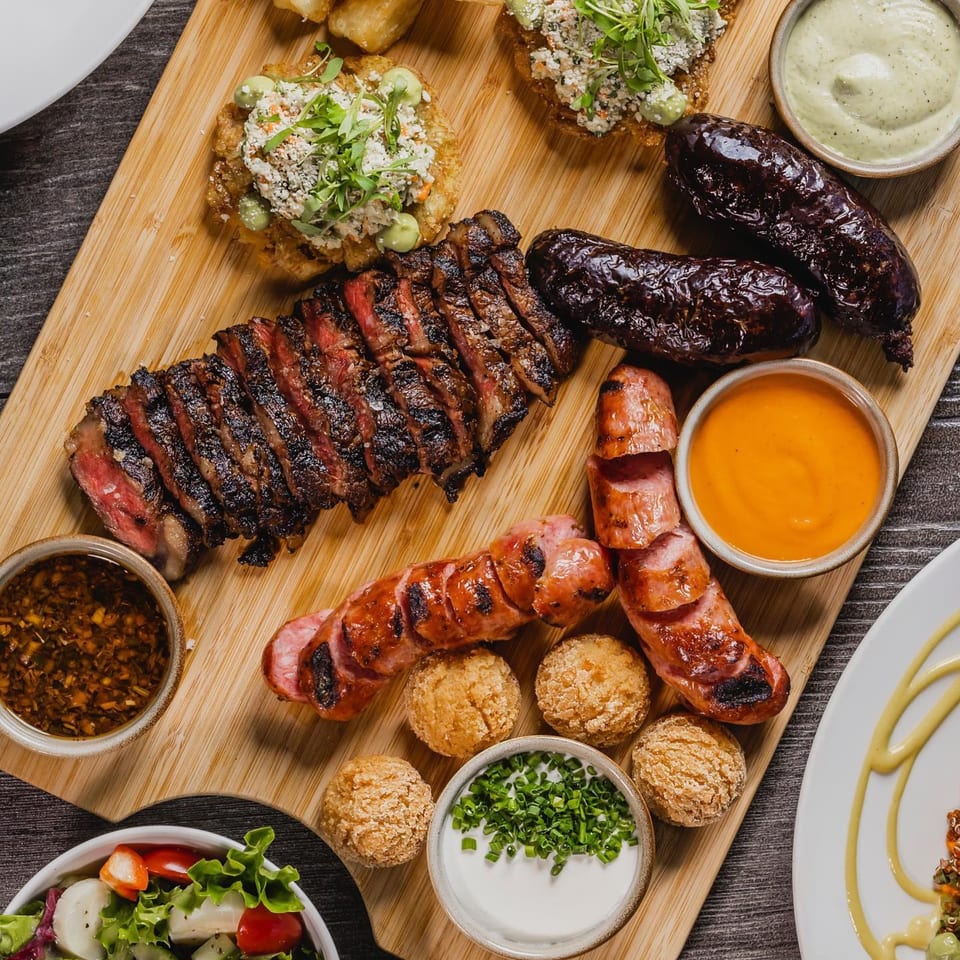 A wooden board with sliced steak, sausages, blood sausage, small fried balls, and various dipping sauces.