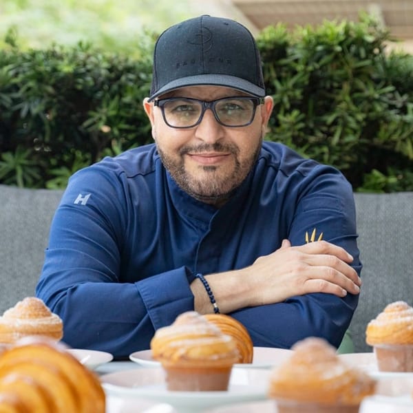 Chef Bachour in a blue chef's coat and cap, smiling behind pastries on a table.