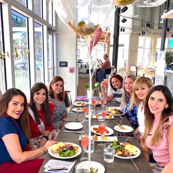 Seven women enjoying brunch with food and pink drinks at a long table in Bachour bakery.