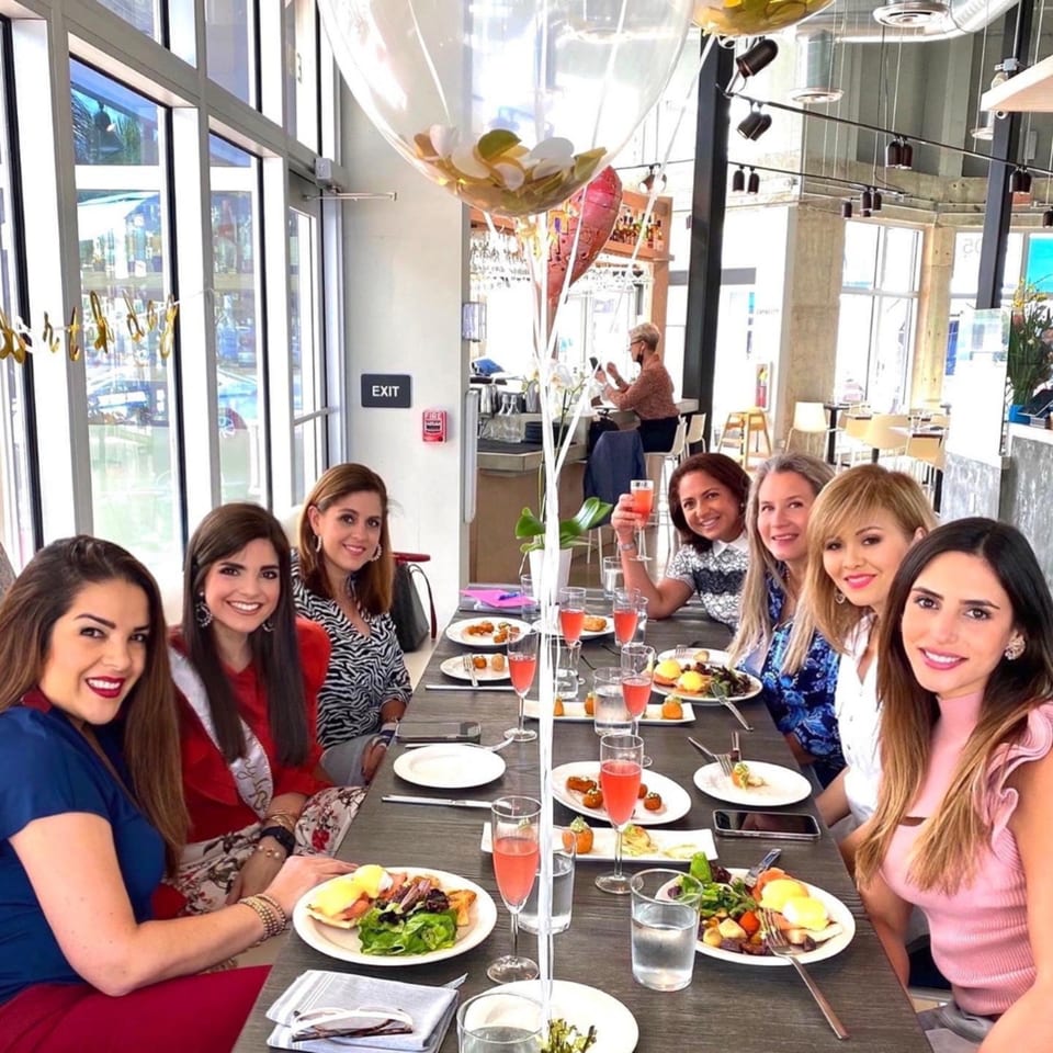 Seven women enjoying brunch with food and pink drinks at a long table in Bachour bakery.