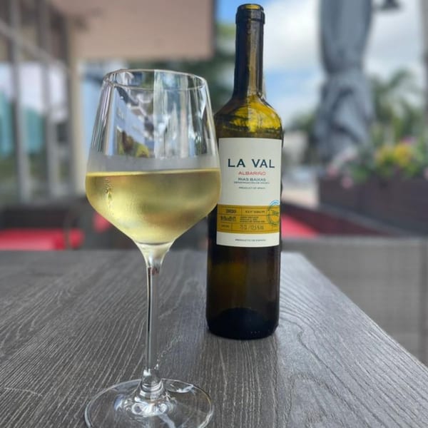A glass of white wine and a bottle of La Val Albarino on a wooden table outdoors.