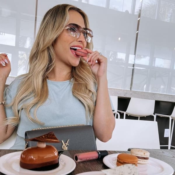 A woman in sunglasses bites into a pink macaron at Bachour, with pastries and macarons on the table.