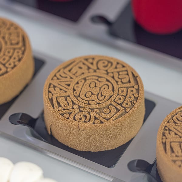Close-up of a round, brown Bachour dessert with intricate patterns on top, on a black tray.
